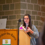 Maela Panei présente son travail de bachelor lors de l'AG 2024 de Biblioromandie. Elle se tient derrière un pupitre portant l'écusson d'Yverdon-les-Bains et tient un livre ouvert.