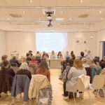 Vue de l’assemblée générale dans la salle Alfred Gründwald de la Médiathèque Valais à Brigue le 4 avril 2025. Le comité se tient sur scène devant une salle dans laquelle on voit le public assis, de dos. Sur scène devant gauche à droite : Lara Jovignot, Fanny Béguelin, Michel Gorin, Julie Greub, Elsa Carloni, Sven Vuillamy, Sylvie Béguelin, Bénoît Epron