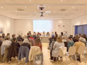 Vue de l’assemblée générale dans la salle Alfred Gründwald de la Médiathèque Valais à Brigue le 4 avril 2025. Le comité se tient sur scène devant une salle dans laquelle on voit le public assis, de dos. Sur scène devant gauche à droite : Lara Jovignot, Fanny Béguelin, Michel Gorin, Julie Greub, Elsa Carloni, Sven Vuillamy, Sylvie Béguelin, Bénoît Epron