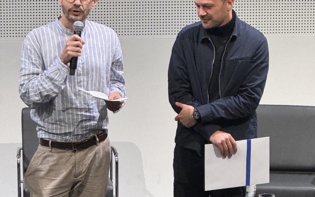 Mathieu Cevey, président de l'AGBD, remet le Prix romand en science de l'information 2025 à Stephen Vallot, sur la scène de l'auditorium de la Haute école de Gestion de Genève, Carouge, le 19 novembre 2025. La photo montre les deux hommes côte à côte en plan pied. Mathieu Cevey sur la gauche de l'image tient un micro et lit et prononce un discours. Stephen Vallot sur la droite de l'image regarde dans sa direction.