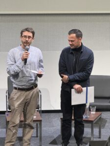 Mathieu Cevey, président de l'AGBD, remet le Prix romand en science de l'information 2025 à Stephen Vallot, sur la scène de l'auditorium de la Haute école de Gestion de Genève, Carouge, le 19 novembre 2025. La photo montre les deux hommes côte à côte en plan pied. Mathieu Cevey sur la gauche de l'image tient un micro et lit et prononce un discours. Stephen Vallot sur la droite de l'image regarde dans sa direction.