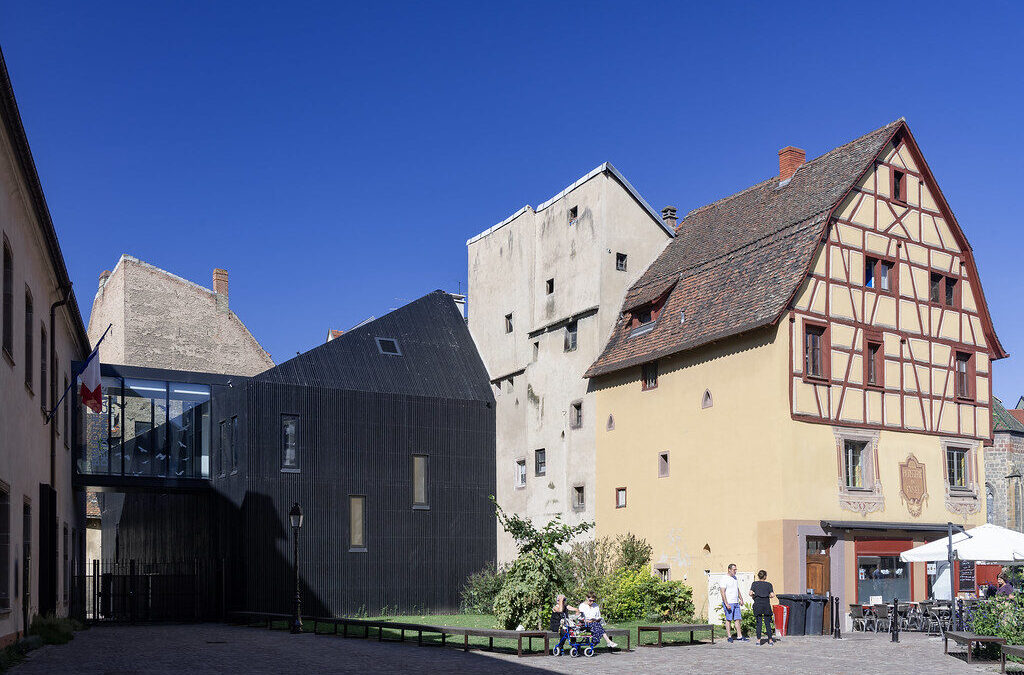 Vue extérieure de la Bibliothèque des Dominicains à Colmar. On voit plusieurs bâtiments sur la photo. Tout à gauche un bâtiment d'un étage, en longueur, vu de perspective. Un drapeau français est placé sur la façade. Dans l'ombre, sur sa droite, relié par une galerie vitrée moderne à l'étage et jusqu'au centre de l'image, un petit immeuble moderne entièrement noir avec des fenêtres étroites et haut de 3 étages. Il est collé sur la droit à un immeuble blanc un peu plus grand, de 5 étages, mais qui semble assez étroit. Enfin sur la droit de l'image, on voit une grande maison à colombage de 5 étage, qui semble assez typique de la région. Il y une terrasse de restaurant devant cette maison, sur une petite place. Le ciel est bleu, sans nuages. L'ambiance est estival et deux groupes de deux personnes discutent au centre de l'image.