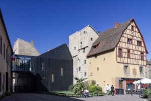 Vue extérieure de la Bibliothèque des Dominicains à Colmar. On voit plusieurs bâtiments sur la photo. Tout à gauche un bâtiment d'un étage, en longueur, vu de perspective. Un drapeau français est placé sur la façade. Dans l'ombre, sur sa droite, relié par une galerie vitrée moderne à l'étage et jusqu'au centre de l'image, un petit immeuble moderne entièrement noir avec des fenêtres étroites et haut de 3 étages. Il est collé sur la droit à un immeuble blanc un peu plus grand, de 5 étages, mais qui semble assez étroit. Enfin sur la droit de l'image, on voit une grande maison à colombage de 5 étage, qui semble assez typique de la région. Il y une terrasse de restaurant devant cette maison, sur une petite place. Le ciel est bleu, sans nuages. L'ambiance est estival et deux groupes de deux personnes discutent au centre de l'image.